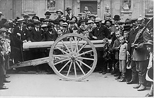 [Crowd of German civilians, including children, surrounding a cannon which has a garland hanging from the muzzle]